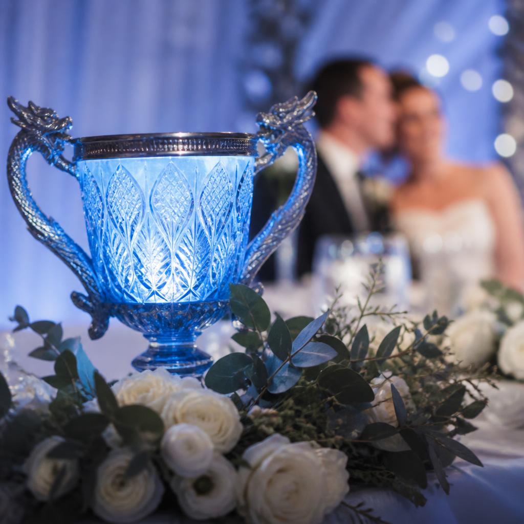 Blau leuchtender Trimagischer Pokal mit Drachenhenkeln im Vordergrund, dahinter unscharf das Brautpaar, umgeben von weißen Rosen und Kerzen